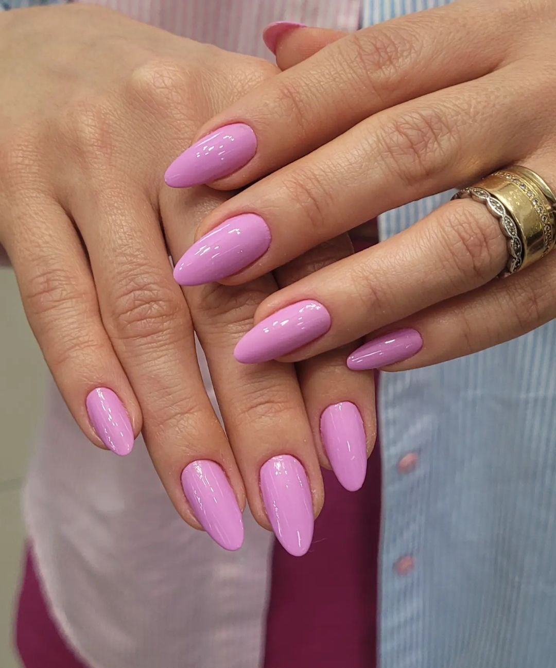 Unhas longas e ovais com esmalte rosa pastel brilhante. A moda destaca cores vibrantes e formas elegantes para unhas. A mão exibe anéis dourados em estilo vintage. Camisa com listras em tons pastel ao fundo.