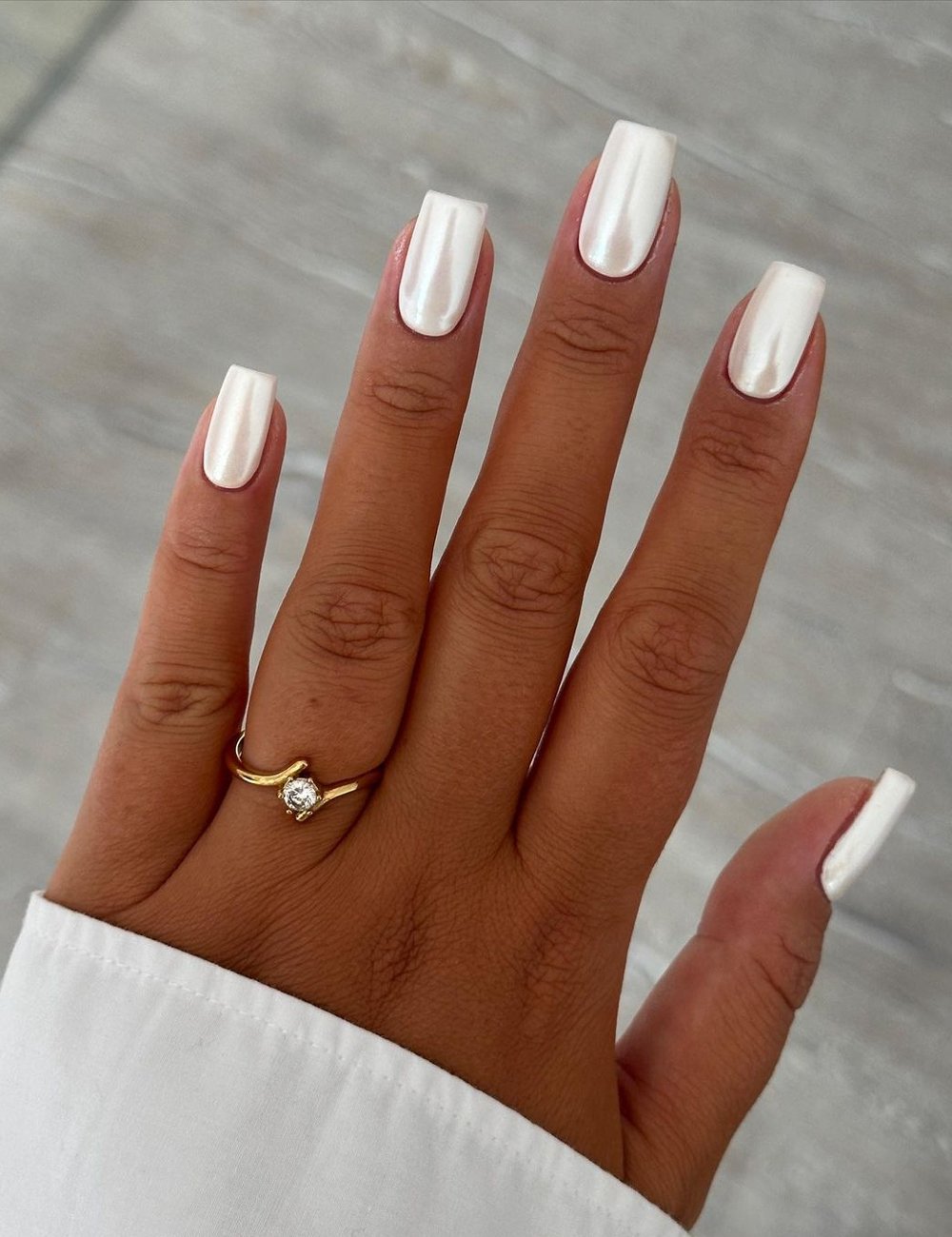 Mão com unhas compridas e formatadas em formato quadrado, pintadas com esmalte branco perolado e top coat. A pessoa usa uma camisa branca e um anel dourado com um pequeno brilho, seguindo um estilo elegante e minimalista.