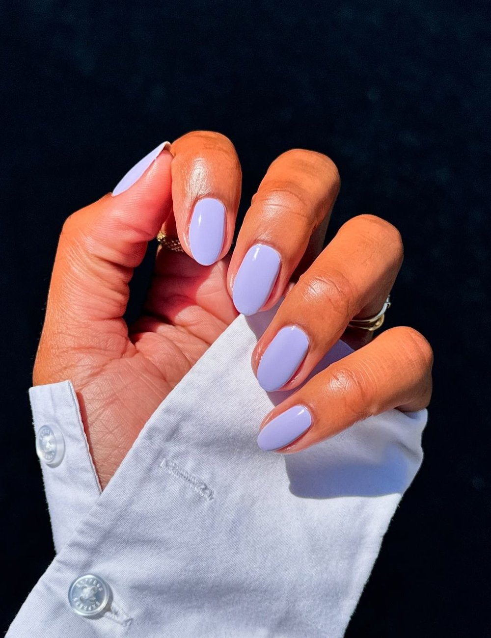 Unhas pintadas em tonalidade lavanda com top coat. A mão veste uma camisa social branca, que contrasta elegantemente com a cor vibrante das unhas. Um anel delicado complementa o visual sofisticado. Ideal para indexação em moda e estilo.