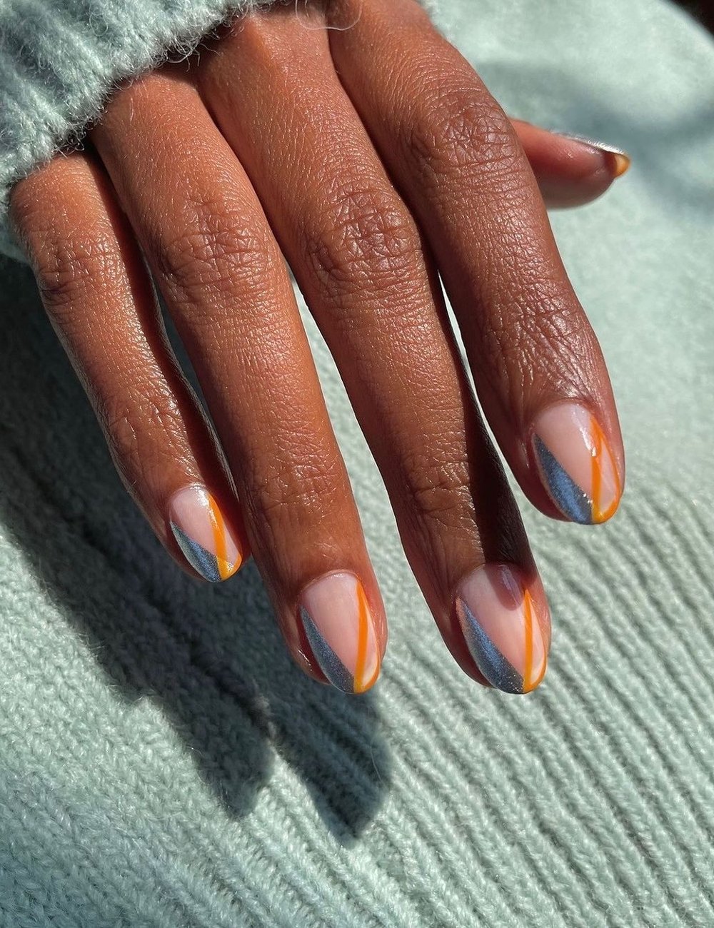 Unhas decoradas artisticamente com listras geométricas de tons laranja e cinza, criando um visual moderno e sofisticado com top coat. A mão repousa sobre um suéter de tricô verde claro, adicionando um toque de conforto e elegância ao conjunto.