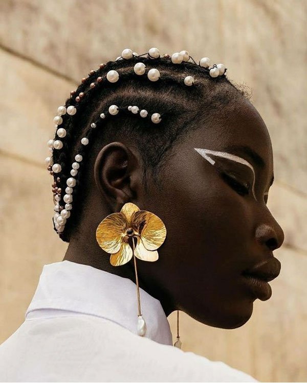 Modelo com tranças adornadas com pérolas, maquiagem de traços brancos sobre os olhos e brinco de flor dourada oversized. Estilo afrocomtemporâneo e elegante.