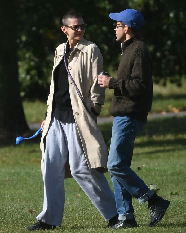 Imagem mostra duas pessoas caminhando ao ar livre. Uma veste trench coat bege, calças de moletom cinza e óculos escuros. A outra usa suéter verde-escuro, boné azul e jeans desbotado. Estilo casual e urbano, valorizando conforto e praticidade.