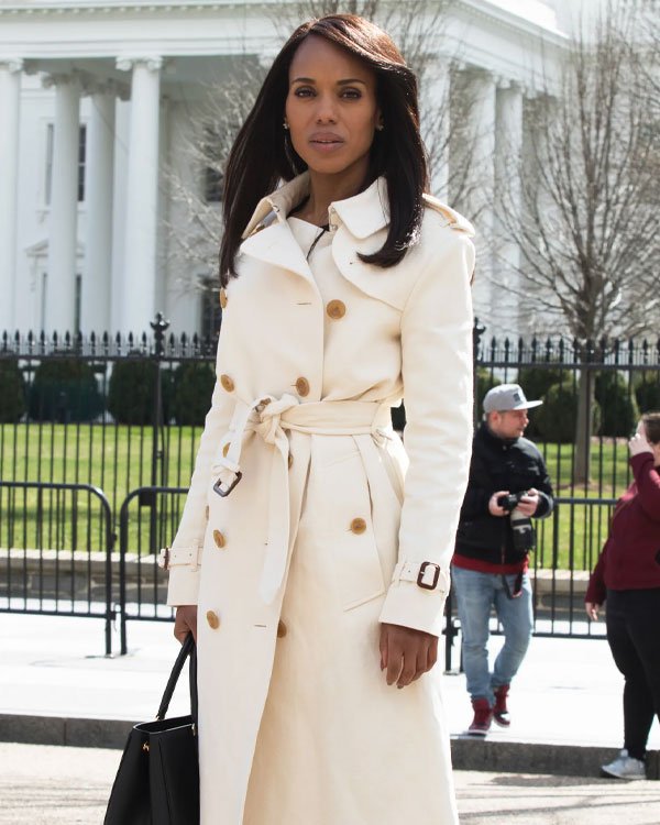 Mulher vestindo um trench coat bege com cinto de amarração e botões grandes, segurando uma bolsa preta. O estilo é clássico e elegante, refletindo uma tendência atemporal da moda outono/inverno. O look é sofisticado e perfeito para ambientes urbanos.