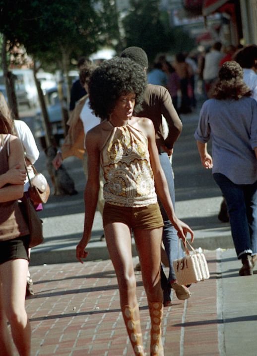 Pessoa caminhando pela rua com visual anos 70: cabelo afro, blusa estampada de amarração no pescoço, shorts curtos de veludo marrom e botas altas com textura. Segura bolsa de alças curta e rígida. Estilo retrô e marcante.