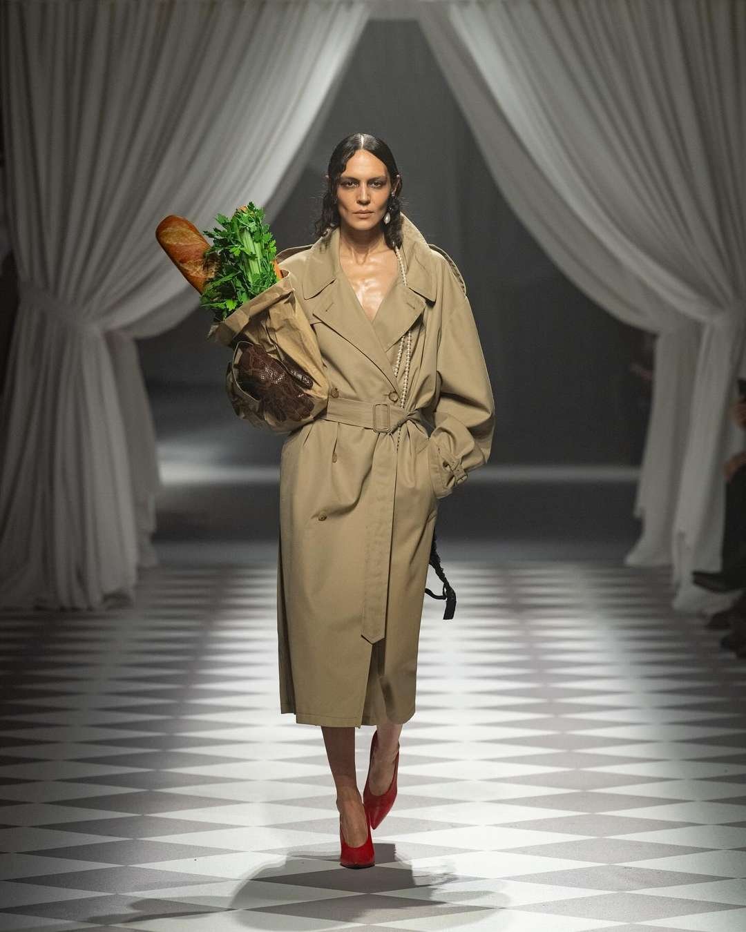 Modelo usando um trench coat bege longo com cinto, carregando uma sacola de papel com baguete e verduras. Nos pés, destaque para sapatos de salto vermelho. A combinação remete a tendências de moda outonal e estilo casual elegante.