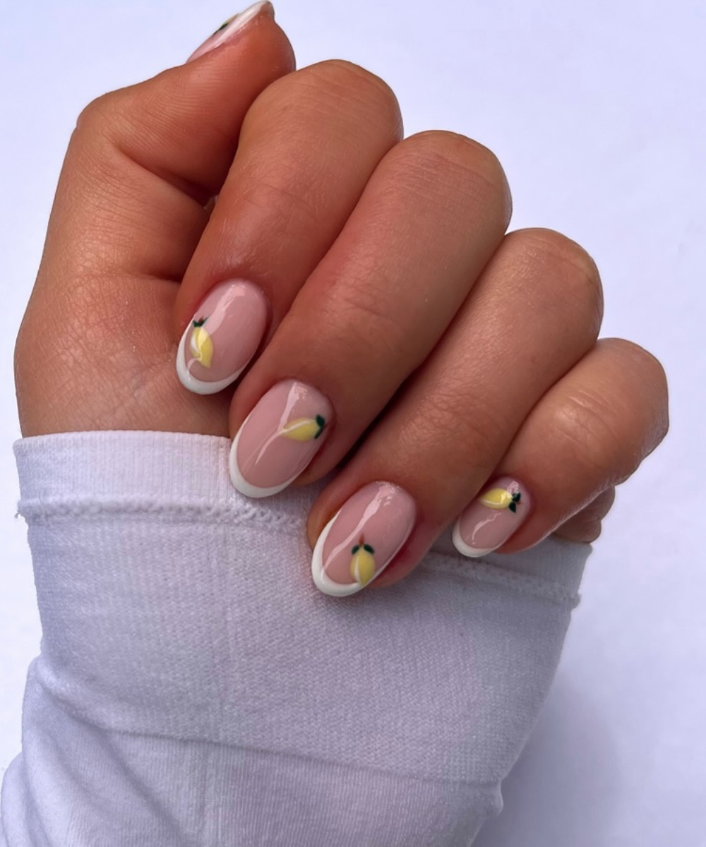 Unhas decoradas com esmalte nude, detalhes de francesinha branca e desenhos de limões pequenos. Tendência de nail art delicada e sofisticada. Mão com blusa branca de manga longa, destacando estilo minimalista e elegante.