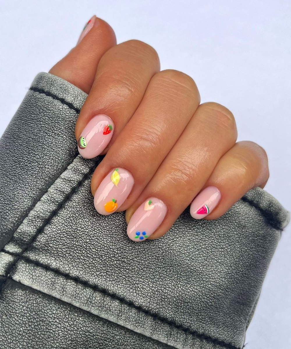 Unhas curtas e arredondadas com esmalte rosa claro, decoradas com desenhos coloridos de frutas e flores, em evidência de tendência de nail art divertida e descontraída. Jaqueta cinza com textura, remetendo a estilo urbano e casual.