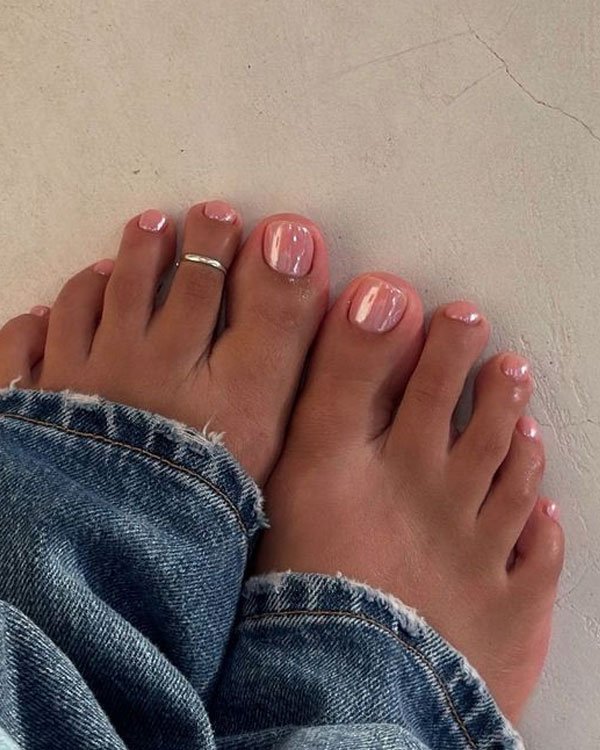 Unhas dos pés pintadas com esmalte rosa claro, estilo natural, e anel de prata no segundo dedo. Os pés estão trajando calça jeans com a barra desfiada, seguindo a tendência de moda casual e descontraída.