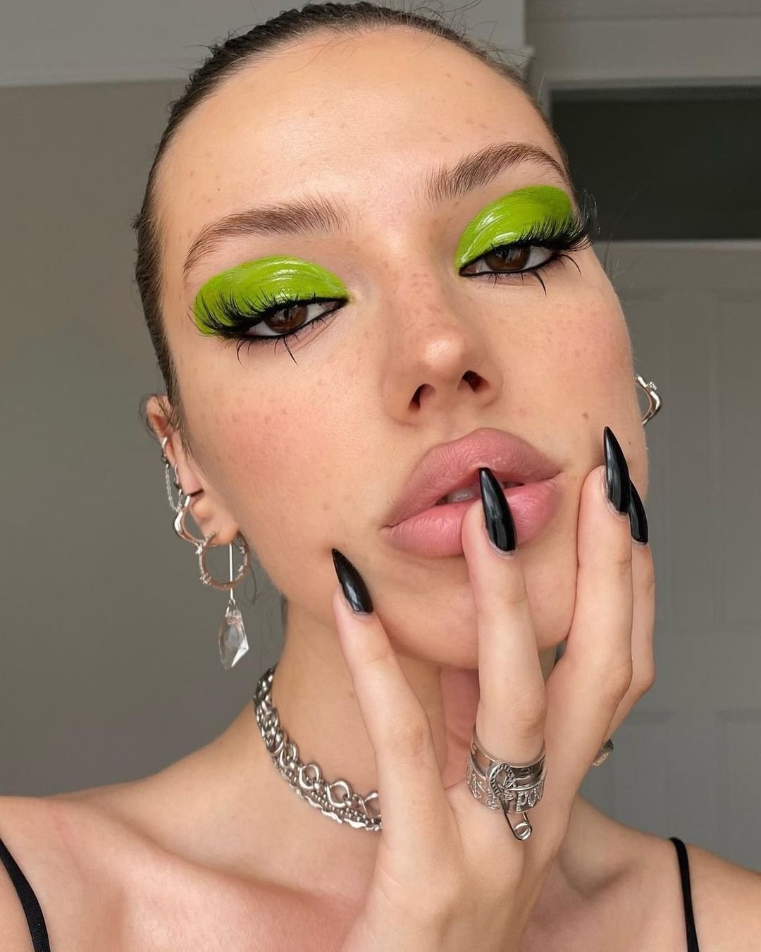 Maquiagem ousada com sombra verde neon e cílios volumosos, unhas longas e pretas estilo stiletto, acessórios prateados, incluindo brincos grandes e anéis, além de gargantilha de corrente prateada. Visual moderno e arrojado.