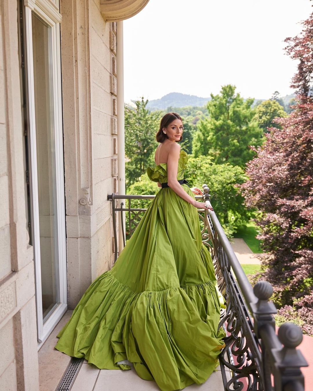 Mulher vestindo um elegante vestido longo verde com babados no decote, posando em uma varanda. O look é sofisticado e segue tendências de moda com foco em cores vibrantes e design volumoso, ideal para eventos formais e ocasiões especiais.