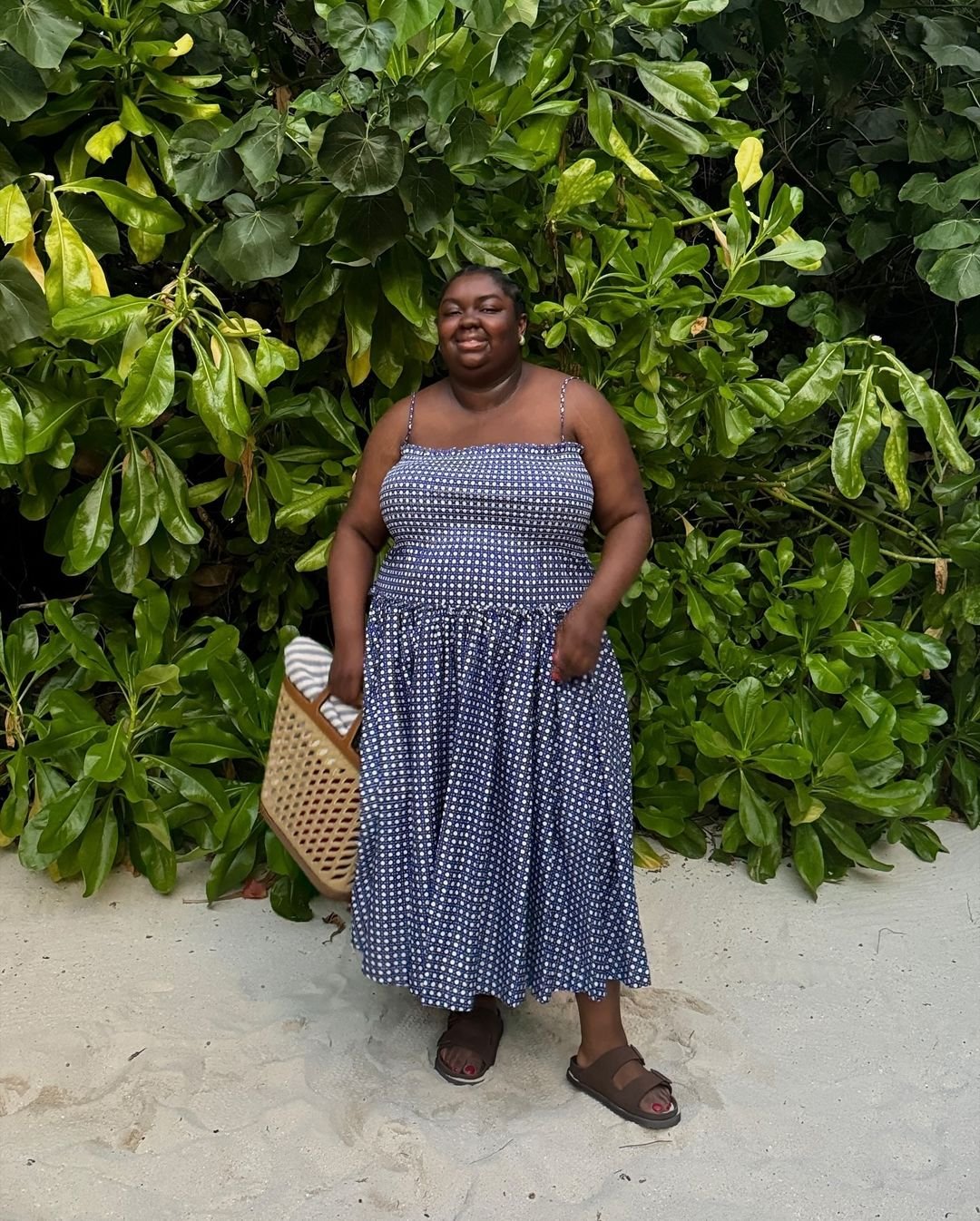 Pessoa vestida com um vestido de alças finas azul-marinho com estampa branca de bolinhas, estilo casual e confortável. Completa o look com sandálias marrons e uma bolsa de palha, criando um visual descontraído perfeito para ambientes de praia.