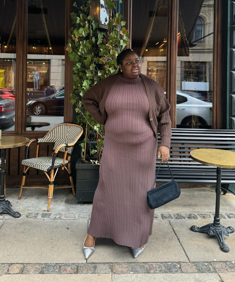 Mulher posa em frente a um café, vestindo um longo vestido canelado marrom, acompanhado de um cardigã marron sobreposto. Completa o look com sapatilhas prateadas de bico fino e uma bolsa preta pequena. Estilo sofisticado e atual.