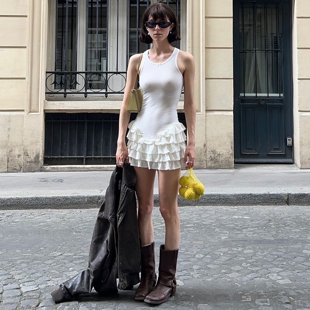 Pessoa vestida com estilo urbano-chic. Usa vestido branco justo com babados na saia, óculos de sol pretos, botas marrons e carrega uma jaqueta marrom e uma bolsa amarela. Tendência mescla delicadeza do vestido com robustez das botas e jaqueta.