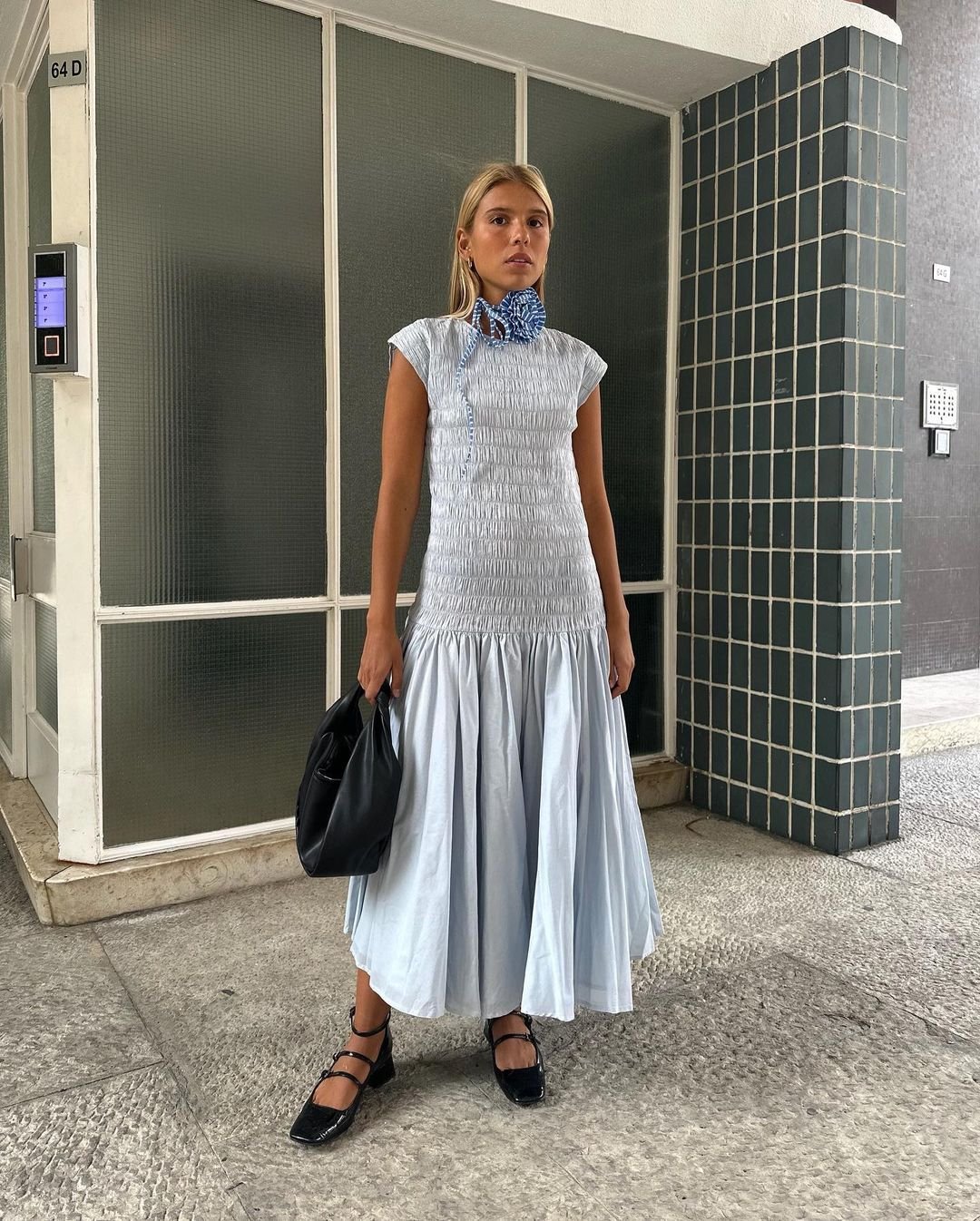 Mulher vestindo um vestido midi azul claro com detalhes franzidos e mangas curtas. Ela está com um lenço azul no pescoço, segurando uma bolsa preta e usando sapatilhas pretas com tiras. Visual elegante e moderno, com toques clássicos e casuais.