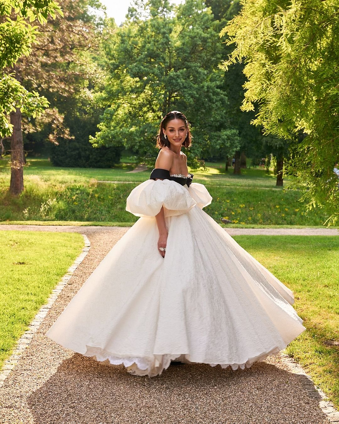 Mulher em vestido extravagante de gala em ambiente ao ar livre. Vestido longo branco com corpete preto ombro a ombro, mangas bufantes e saia volumosa. Estilo elegante e romântico, ideal para eventos formais. Tendência em moda de alta-costura.