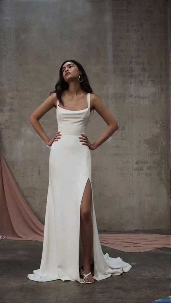 Mulher usando vestido longo branco com fenda na perna, alças finas e cauda ligeira, combinando com sandálias delicadas. A moda expressa elegância minimalista e sofisticação. O fundo neutro contrasta com a simplicidade e beleza do traje.