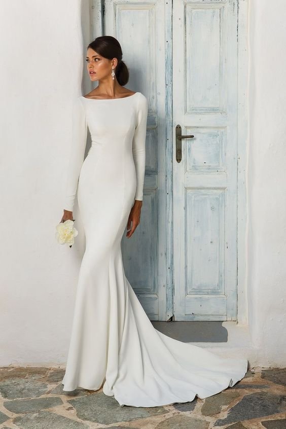 Vestida com um elegante vestido de noiva branco, de manga longa e corte sereia, evidenciando um estilo minimalista e sofisticado. O design simples e atemporal realça a silhueta feminina. A modelo segura um pequeno buquê e usa brincos compridos.