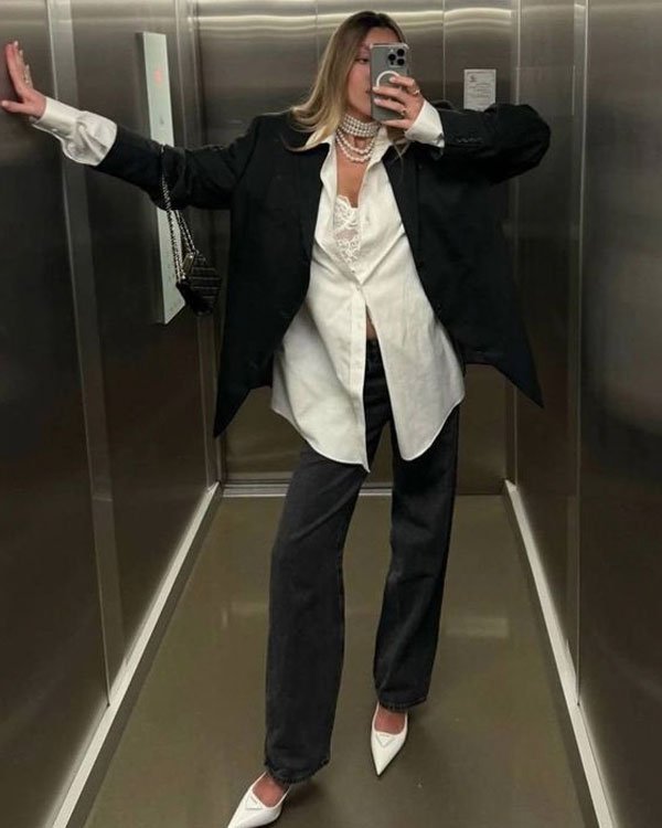 Look moderno com sobreposição de blazer oversized preto, camisa branca larga e renda à mostra. Calça jeans folgada e sapatos de bico fino brancos. Acessório marcante: colar de pérolas e bolsa preta. Trend despojado e sofisticado.