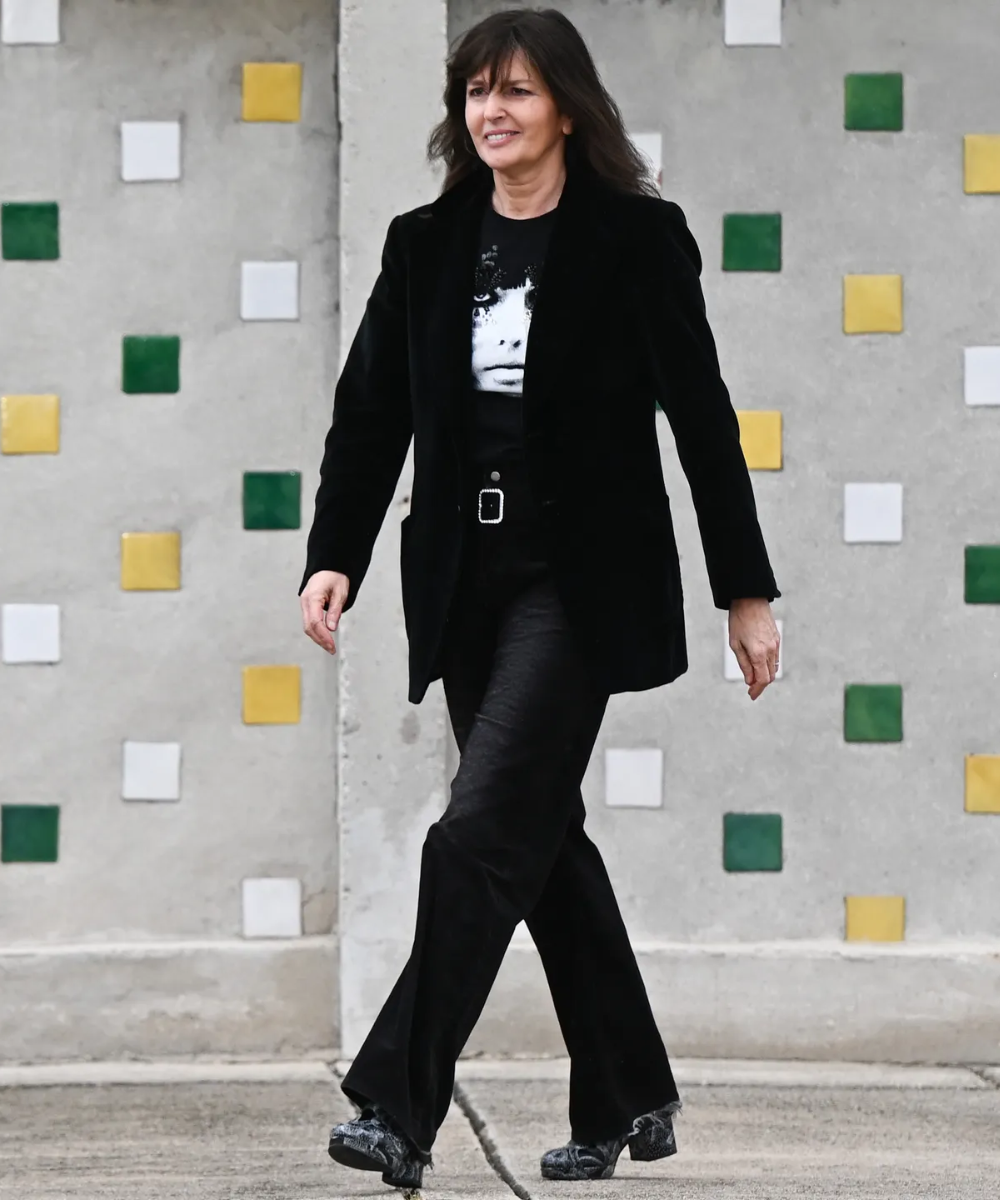 Mulher vestindo blazer preto, camiseta estampada com rosto, calça preta de tecido brilhante e sapatos escuros com salto. Look elegante e moderno, combina peças clássicas com estilo urbano. Tendência: moda monocromática, tecidos texturizados e estampa