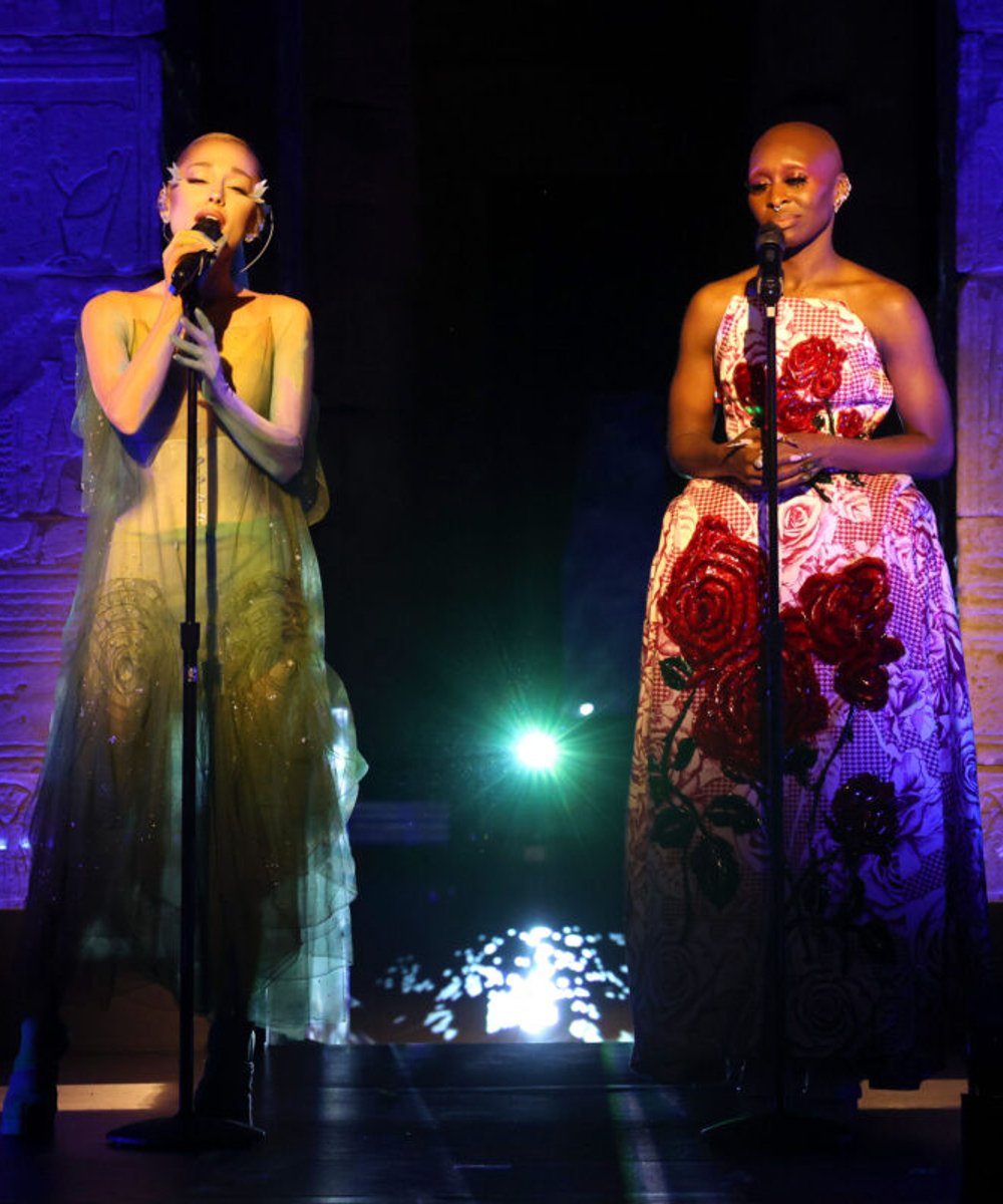 Duas mulheres em palco cantando no microfone. A da esquerda usa um vestido verde translúcido, estilo etéreo, tendência de moda transparente. A da direita veste um vestido estruturado com estampa floral em vermelho, tendência de moda statement. Wicked