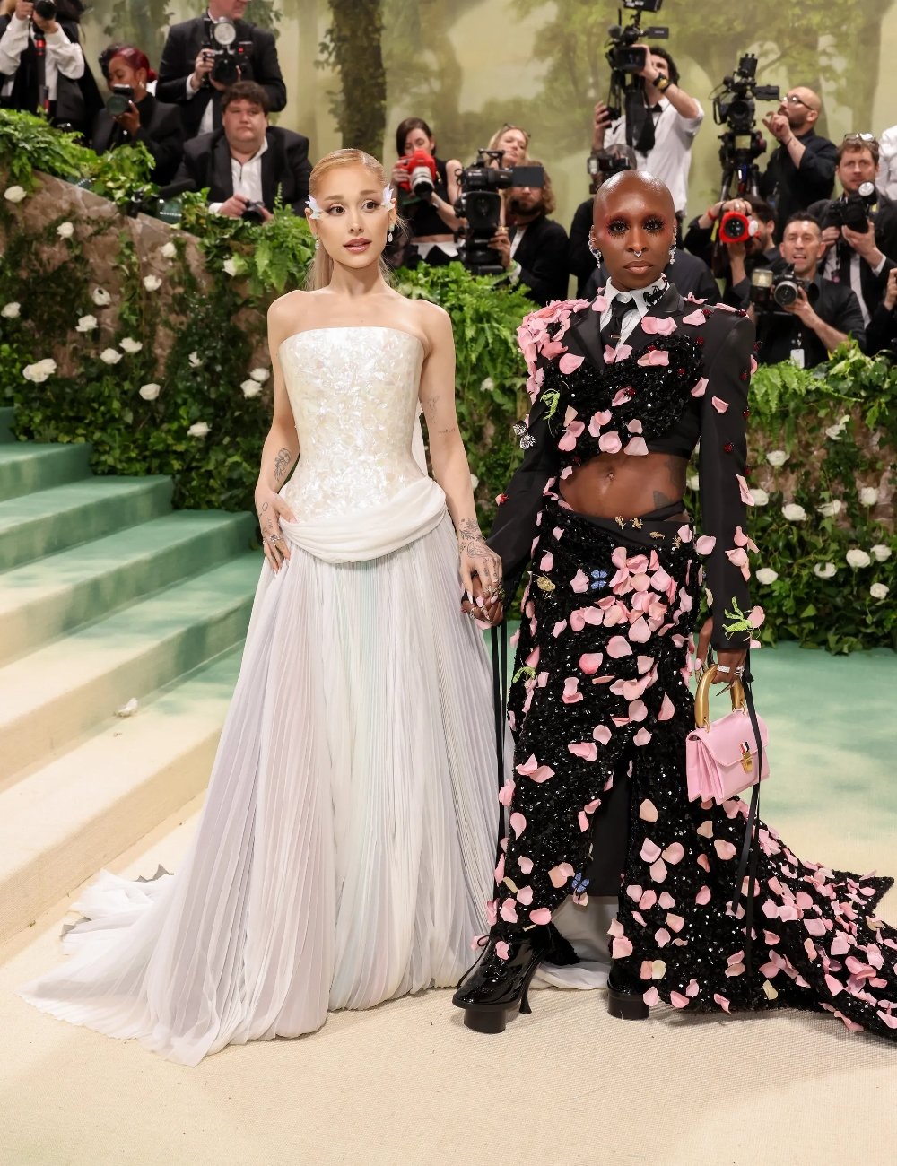 Duas pessoas em um evento de gala: uma usa vestido branco tomara-que-caia com cauda e acessórios delicados, enquanto a outra veste um conjunto preto com flores aplicadas em 3D, exibindo um visual ousado e moderno. Wicked.