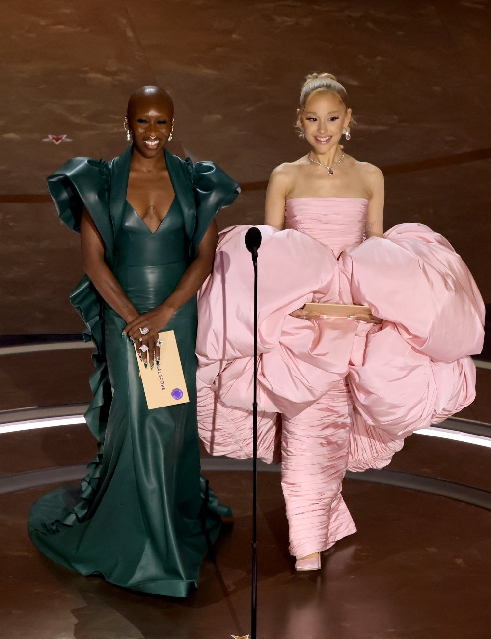 Imagem de duas mulheres em um evento; uma veste um elegante vestido verde com ombros estruturados e a outra usa um volumoso e extravagante vestido rosa. Wicked.