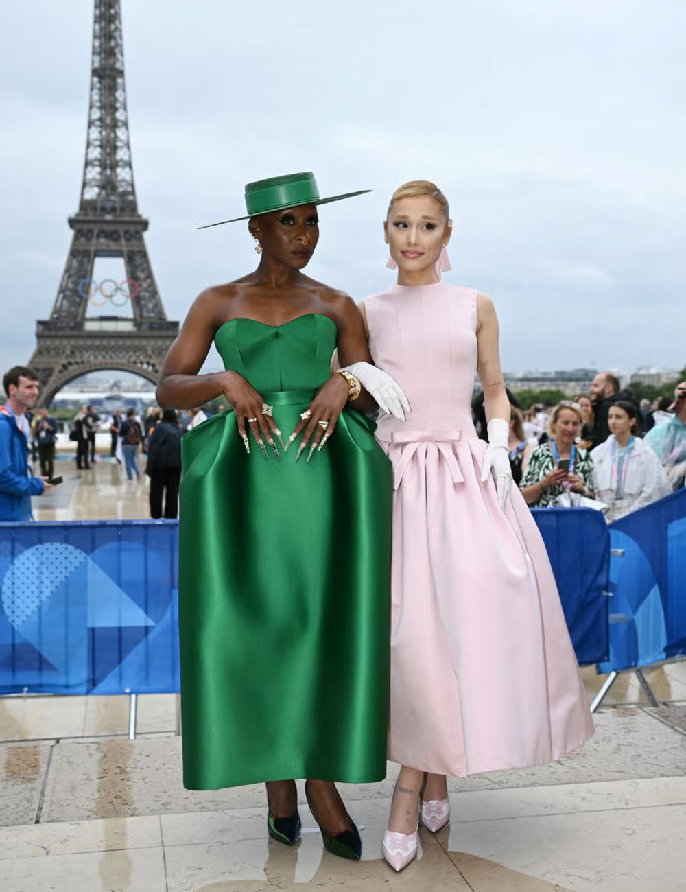 Duas mulheres vestindo roupas elegantes em Paris, com a Torre Eiffel ao fundo. A primeira usa um vestido verde vibrante e chapéu combinando, enquanto a segunda veste um vestido rosa claro e luvas brancas. Wicked.