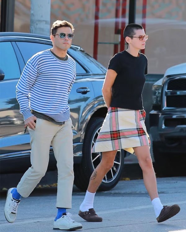 Casal caminhando em rua urbana. Homem veste camiseta listrada azul e branca, calça bege e tênis branco com meias azuis. Mulher usa camiseta preta, saia xadrez e sapatos marrons com meias brancas. Estilos casual e vintage, com influências retrô.