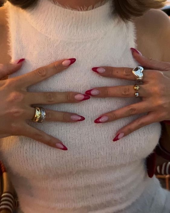 Imagem de uma pessoa vestindo um suéter branco de gola alta. As unhas são longas e decoradas com nail art rosa, com ponta vermelha. Vários anéis modernos adornam os dedos, complementando o visual sofisticado e atual.