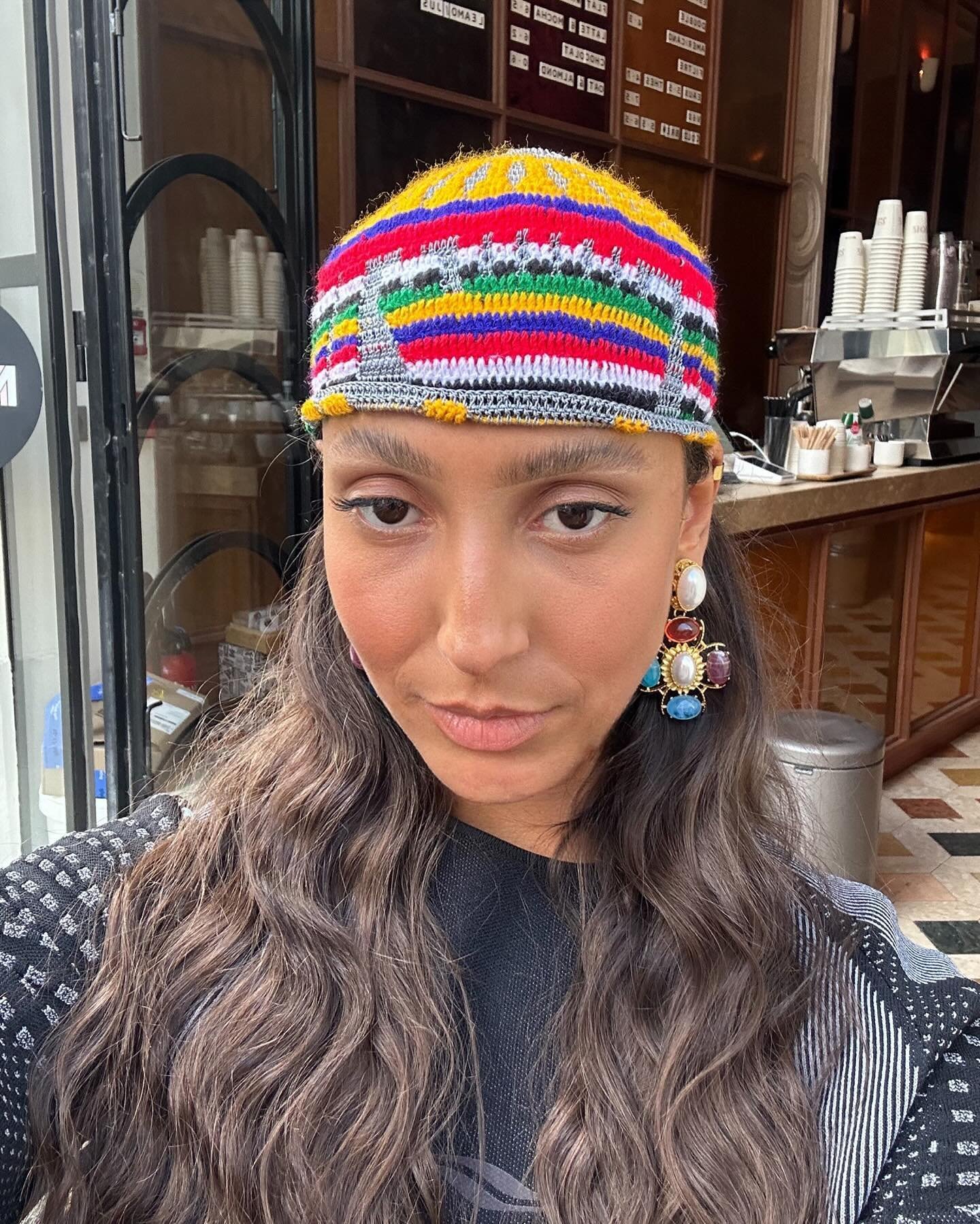 Mulher com gorro colorido de tricô, composto por listras em várias cores. Ela usa brincos pendentes com pedras de diferentes cores e formato solto nos cabelos ondulados. Estilo casual e moderno, com acessórios que destacam um toque boho e criativo.