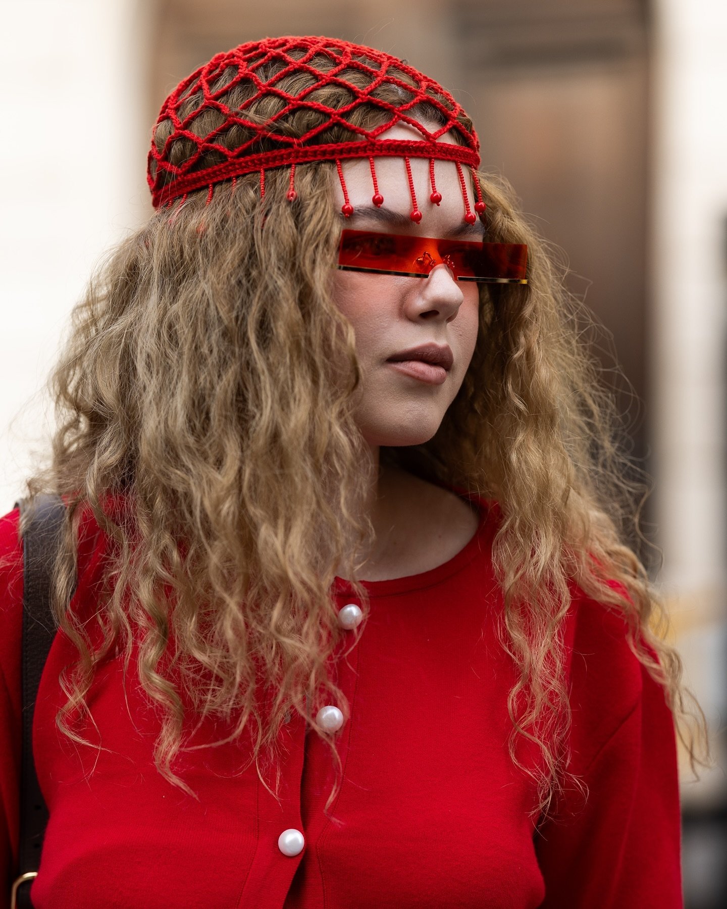 Moda urbana criativa: mulher com chapéu de crochê vermelho, óculos de sol futuristas e blusa vermelha com botões grandes. Look ousado e inovador, destacando tendências de acessórios únicos e cores vibrantes.