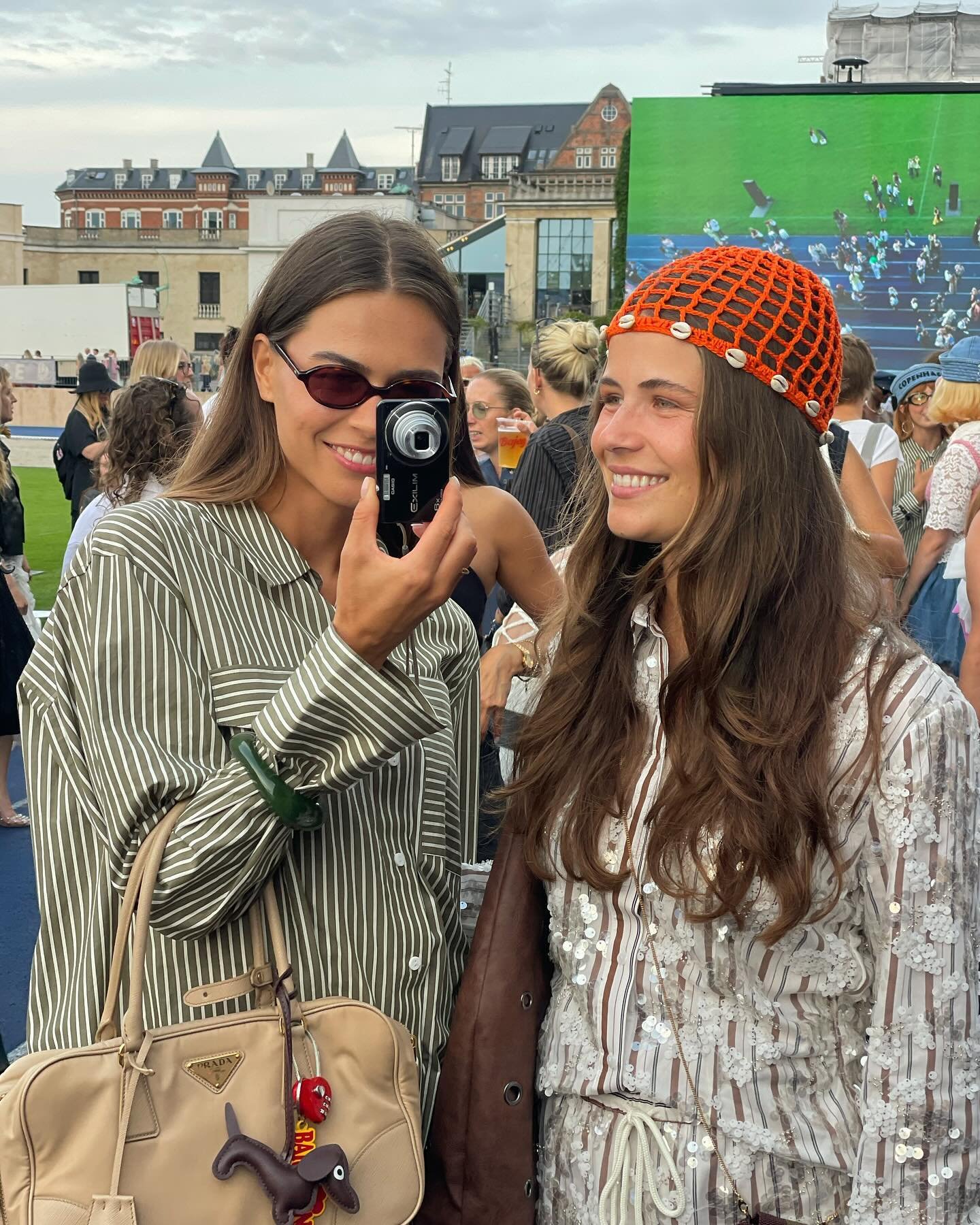 Duas mulheres vestindo tendências fashion: a da esquerda usa camisa listrada verde com óculos escuros e bolsa Prada bege com chaveiro fofo. A da direita veste camisa de lantejoulas e um gorro laranja de crochê com conchas. Estilo casual e moderno.