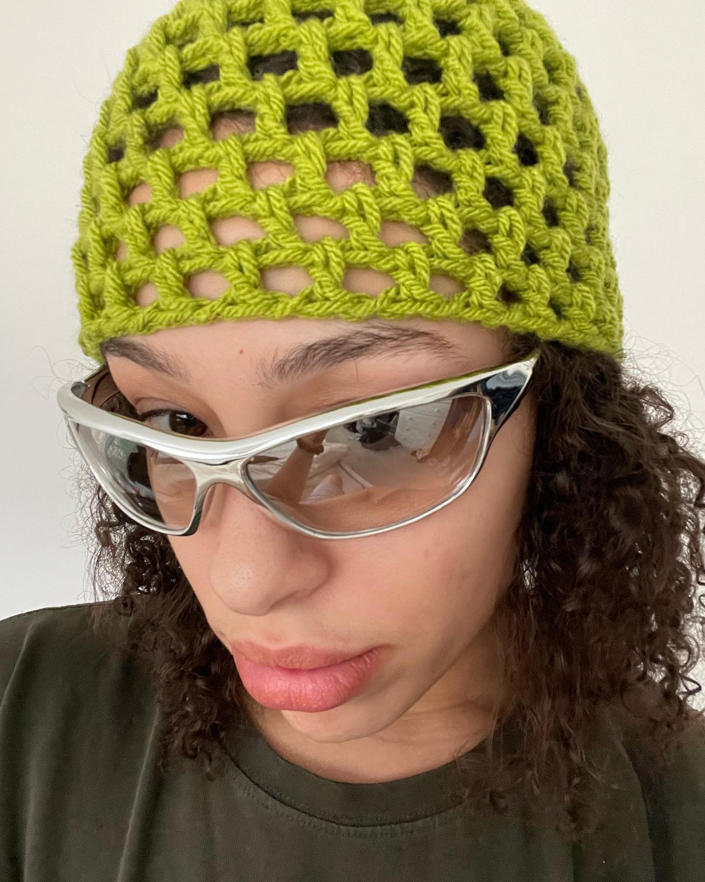 Pessoa usando gorro de crochê verde neon e óculos de sol futuristas com armação prata, vestindo camiseta preta básica. Tendência de moda utilitária e acessório statement, destacando estilo urbano e alternativo.