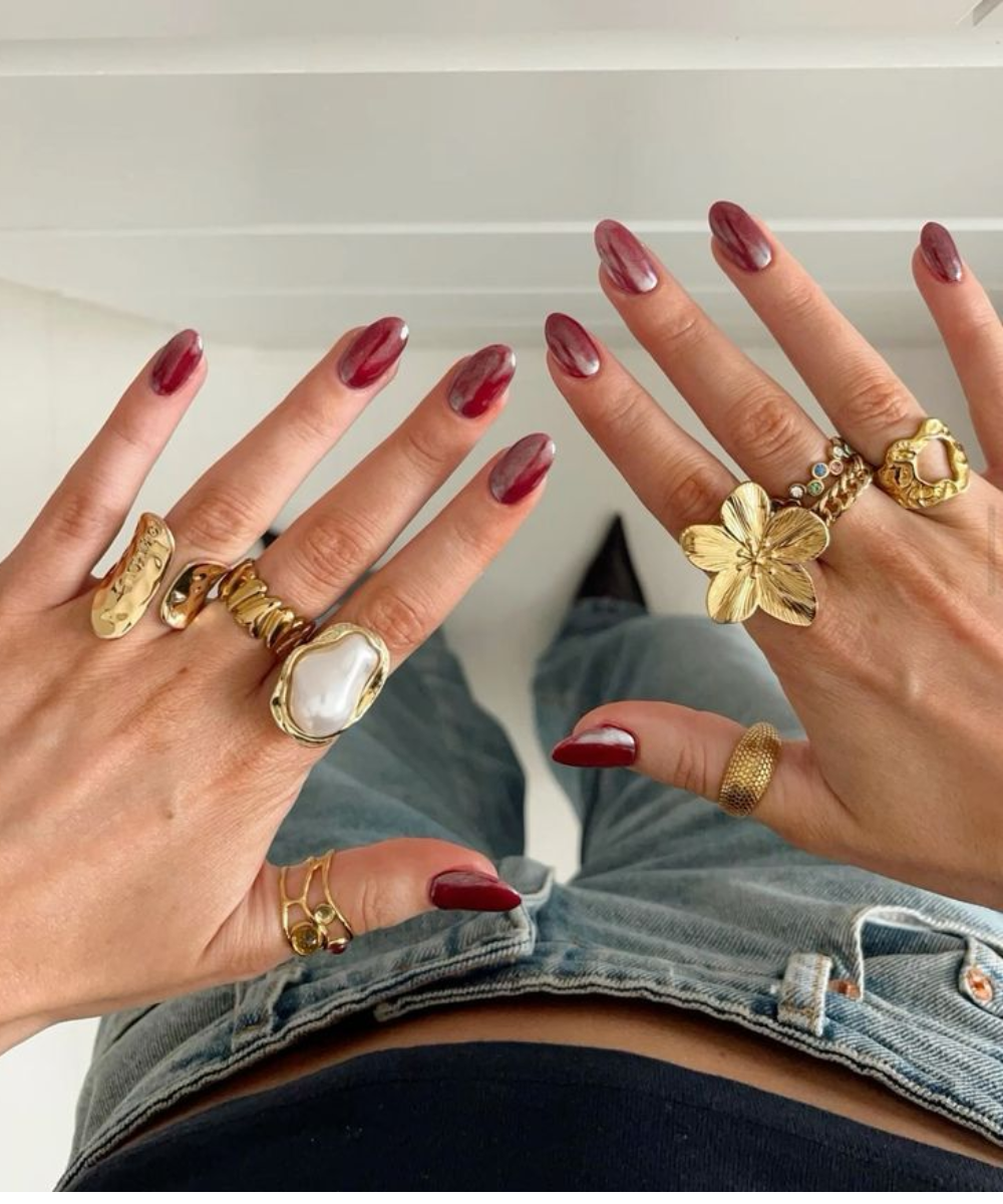 Imagem destaca mãos com unhas pintadas de vermelho e vários acessórios dourados, incluindo anéis grandes e chamativos. A pessoa veste jeans de cintura alta e top preto, refletindo uma mistura de tendências modernas e elegantes.