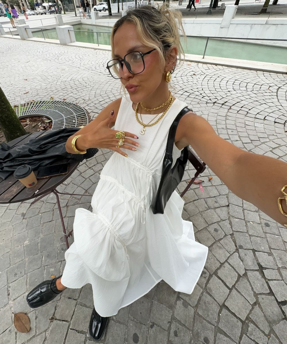 Mulher usando vestido branco longo com textura, óculos grandes, colares dourados, brincos de argola, pulseiras e anéis chamativos. Carregando bolsa preta e calçando sapatos pretos de couro. Estilo moderno e fashionista. Fundo: espaço urbano pavimentado.