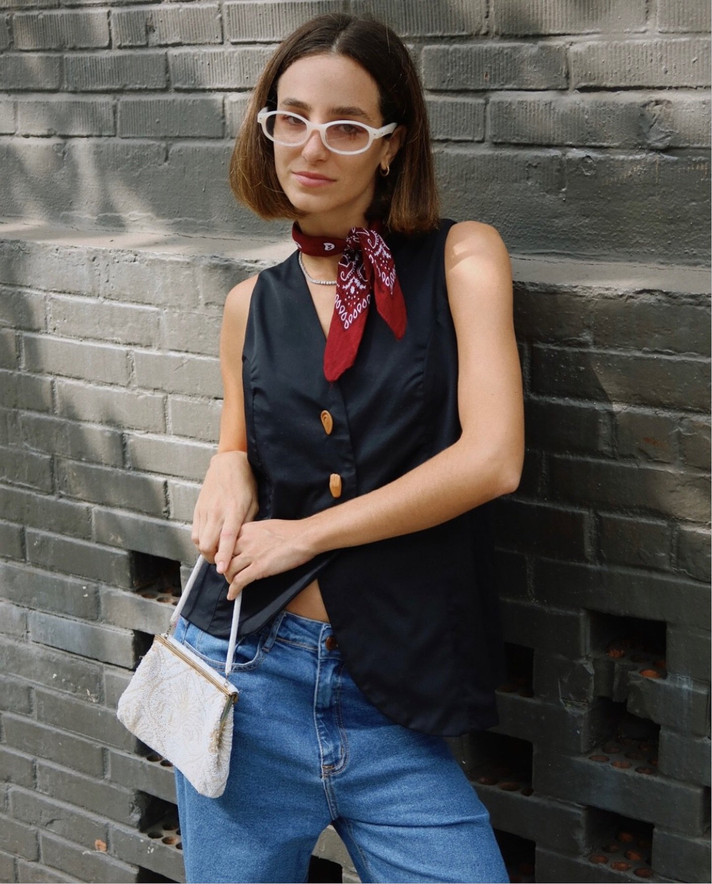 Mulher com óculos de armação branca e lenço vermelho no pescoço, vestindo colete preto com botões, jeans de cintura alta e bolsa de mão branca com bordados. Visual casual com toque vintage e acessórios retrô.