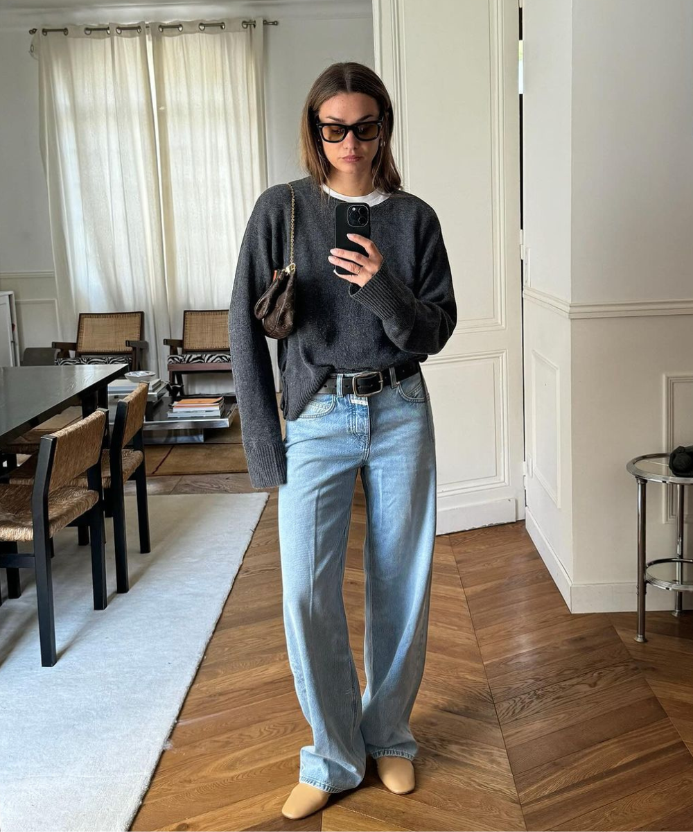 Mulher com visual casual-chique, usando suéter cinza oversized, jeans de cintura alta e corte reto, sapatos bege e óculos escuros grandes. A combinação inclui cinto preto e bolsa marrom. Estilo descontraído e minimalista, tendência em alta no streetw