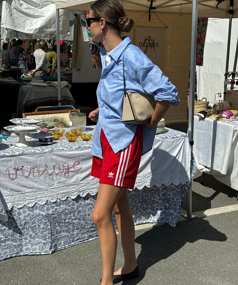 Mulher usando camisa social azul clara, shorts vermelhos Adidas e sapatilhas pretas. Ela carrega uma bolsa bege e usa óculos escuros. O estilo mistura esportivo e casual com toques vintage, ideal para passeios em feiras ou mercados ao ar livre.