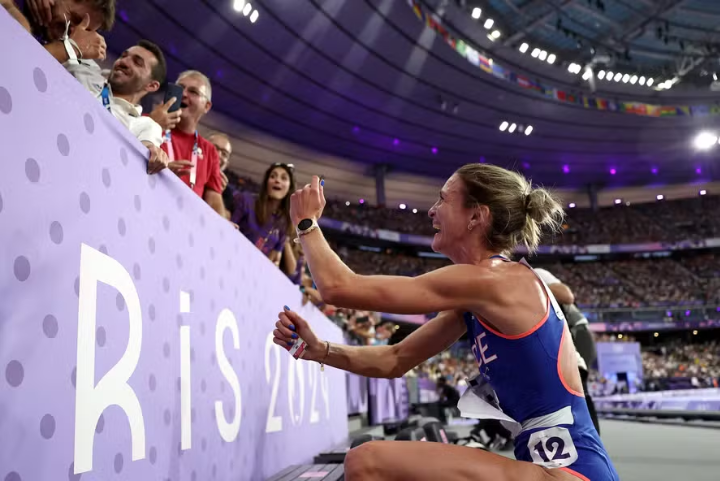 Atleta vestida com uniforme esportivo de competição: maiô azul com detalhes brancos e vermelhos, próprios para alta performance. Cabelo preso em coque. Tendência de moda esportiva focada em funcionalidade e aerodinâmica para competições.