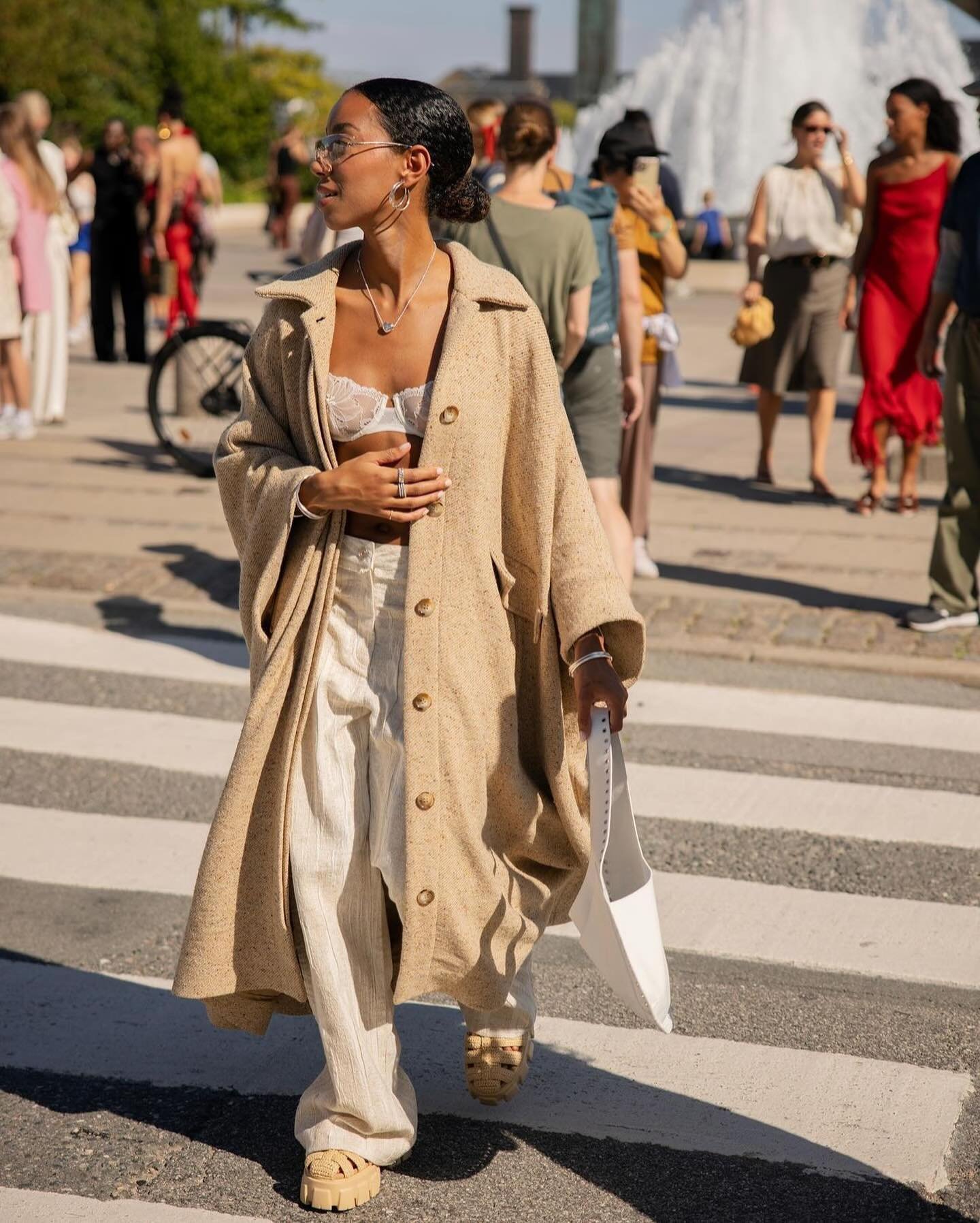 Mulher vestida com um longo casaco bege, calça larga de linho e um top cropped de renda branca. Acessórios incluem óculos de sol, brinco argola, colar e sandálias plataforma bege.