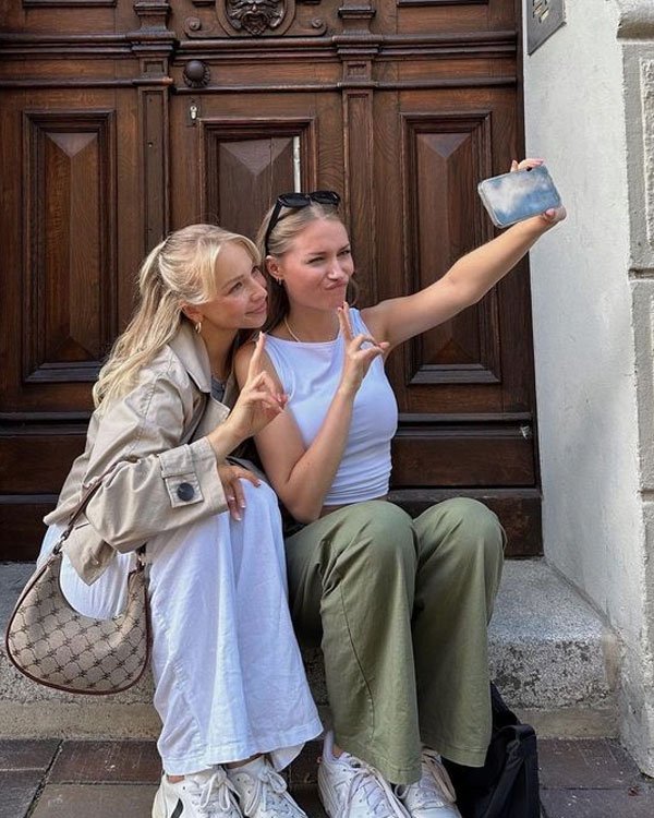 Duas mulheres jovens posando para uma selfie. Moda casual: uma veste trench coat bege, vestido branco, tênis e bolsa pequena estampada, enquanto a outra usa regata branca, calça verde oliva e tênis branco.