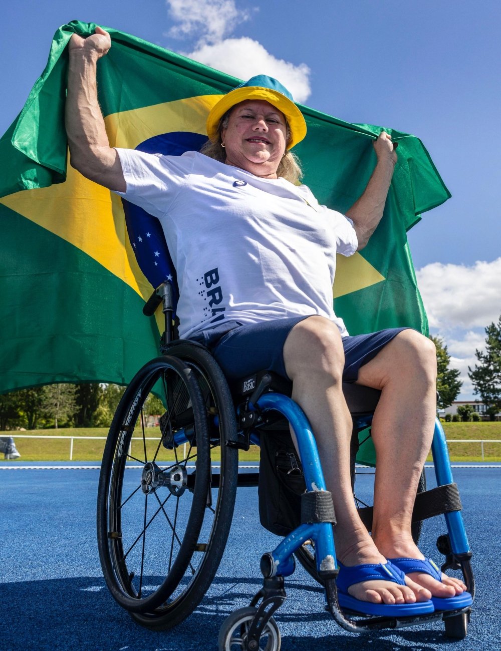 Pessoa em cadeira de rodas vestindo camiseta branca com estampas do Brasil e shorts azul marinho. Acessórios incluem chapéu amarelo e sandálias azuis. Paralimpíadas de Paris 2024.