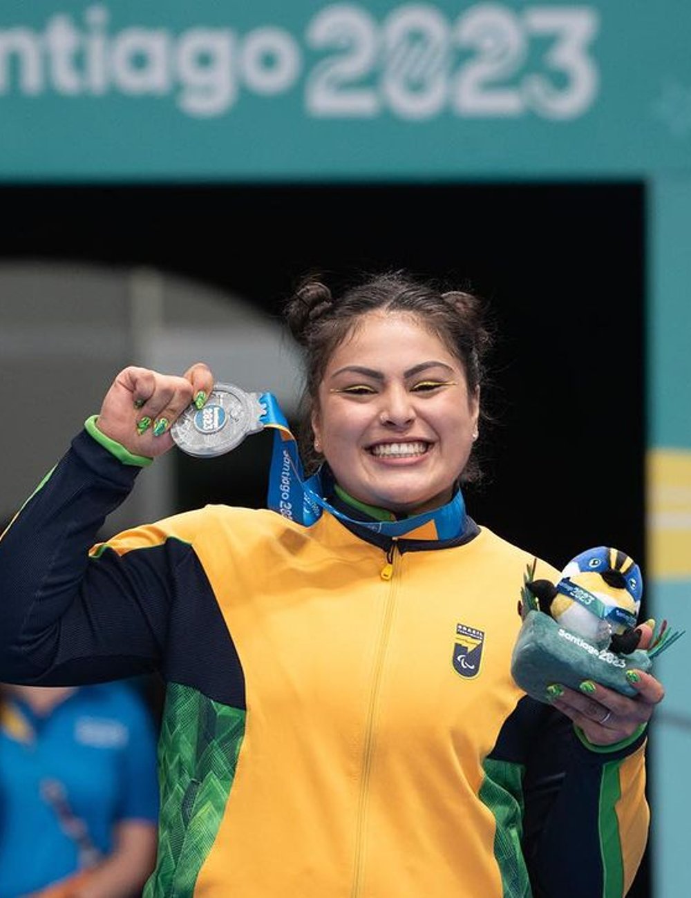 Atleta vestindo uniforme esportivo nas cores verde e amarelo. Roupa inclui jaqueta com zíper e logotipo. Cabelos presos em coques duplos, maquiagem com delineado amarelo. Paralimpíadas de Paris 2024.