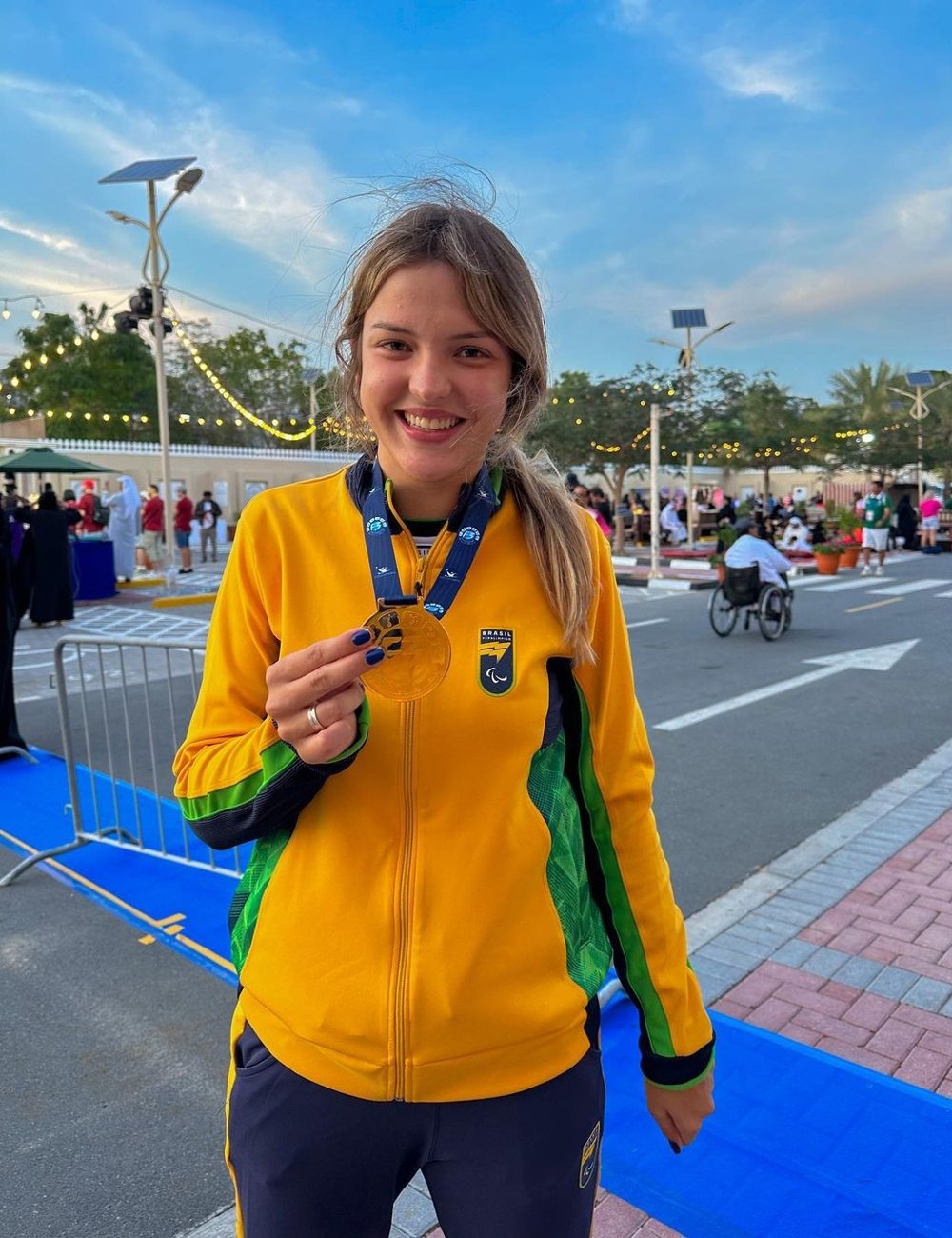 Atleta vestindo uniforme esportivo brasileiro em cores vibrantes de amarelo e verde, exibindo uma medalha de ouro. A jaqueta possui detalhes verdes nas laterais e ziper frontal, combinada com calça esportiva azul escura. Paralimpíadas Paris 2024.