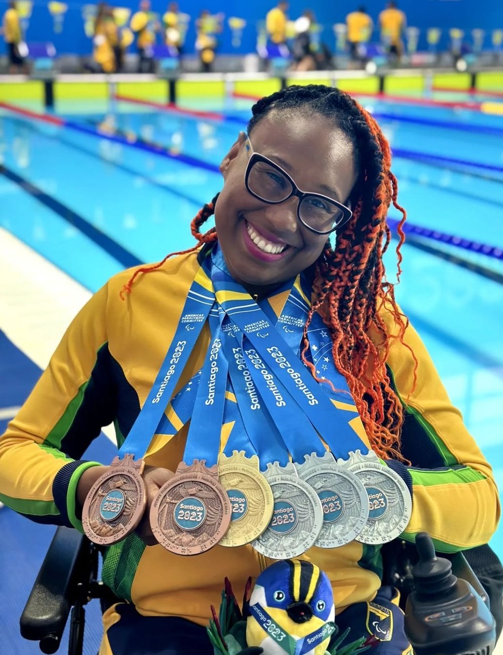 Atleta vestida com agasalho amarelo e detalhes verdes, exibindo múltiplas medalhas ao redor do pescoço. Tranças vermelhas e pretas conferem um estilo moderno e vibrante. óculos completam o look esportivo. Paralimpíadas de Paris 2024.