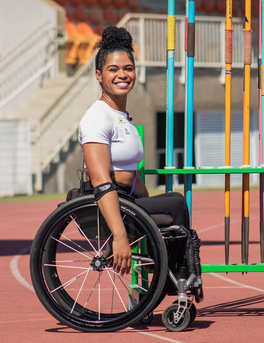 Atleta feminina em cadeira de rodas, vestindo cropped branco justo e calça legging preta, em uma pista de atletismo com dardos ao fundo. Paralimpíadas de Paris 2024.