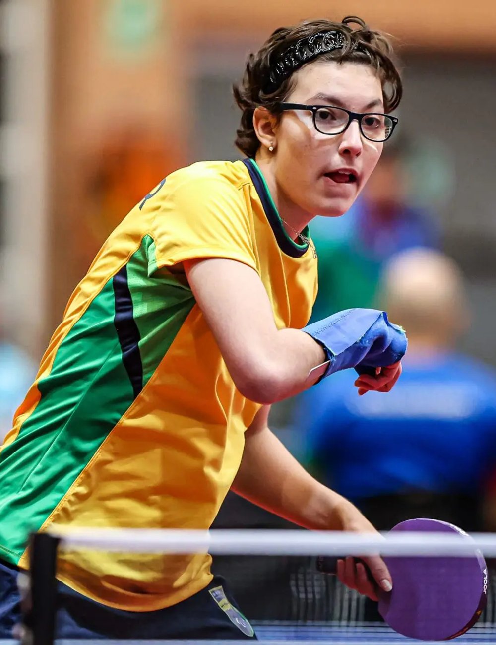 Jovem vestindo camiseta esportiva, predominantemente amarela com detalhes verdes. Usa óculos de aro preto e bandana preta no cabelo curto. Luva esportiva azul na mão esquerda. Paralimpíadas de Paris 2024.