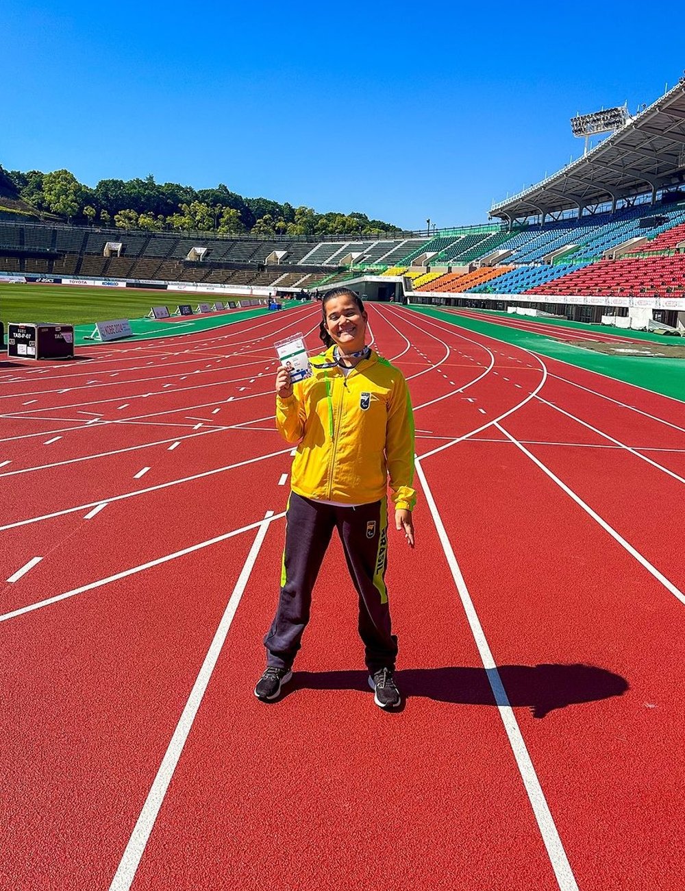 Atleta em pista de atletismo, usando jaqueta esportiva amarela e calça de moletom azul marinho com detalhes verdes. Look casual esportivo, ideal para treinos ao ar livre. Paralimpíadas de Paris 2024