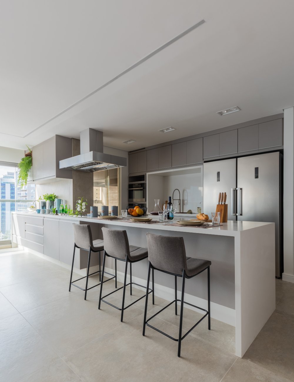 Cozinha moderna com design minimalista, destacando armários embutidos em tons de cinza, banquetas de quartzo branco e eletrodomésticos inox. Ilha central acompanha banquetas estofadas, combinando estética contemporânea e funcionalidade.