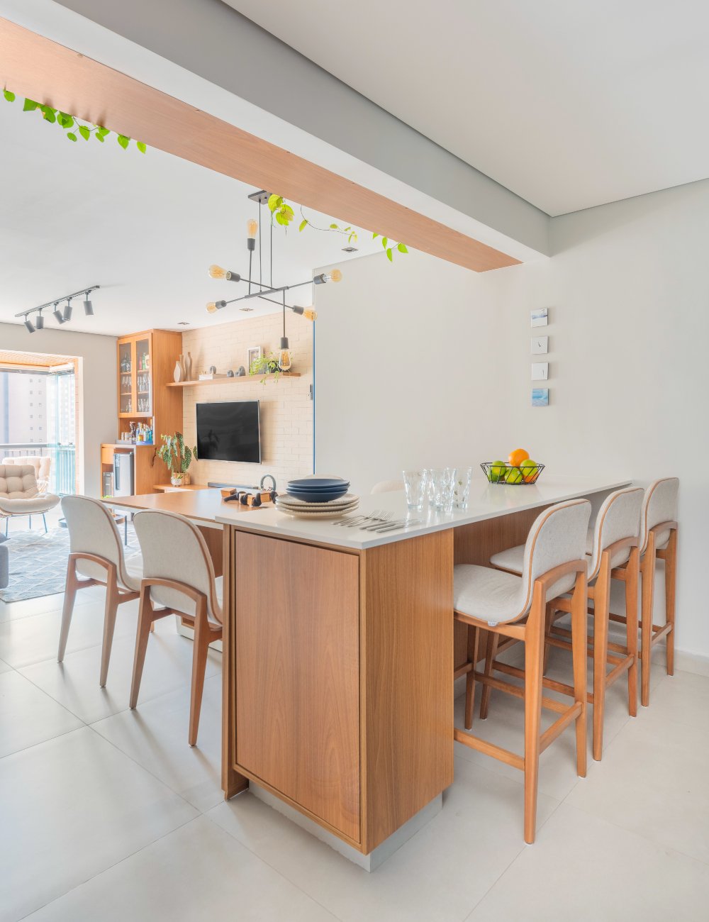 Cozinha moderna com ilha central em madeira e banquetas claras, cadeiras estofadas bege, iluminação pendente preta, e detalhes decorativos com plantas. Estilo contemporâneo com tendência minimalista e uso de materiais naturais.